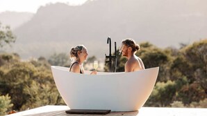 Two people in a large white bathtub outdoors with a mountainous backdrop. - Tiny Aura. (Coxs Crown)