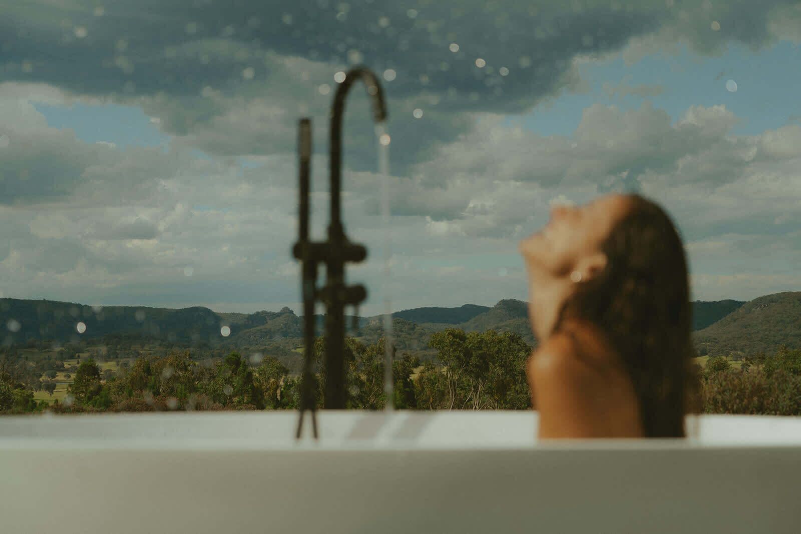 A person in a bathtub with a view of a landscape and mountains in the background.