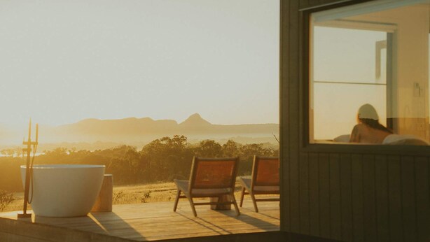 Terraza de madera con una bañera blanca grande, dos sillas de madera y vistas a montañas distantes a través de una ventana.