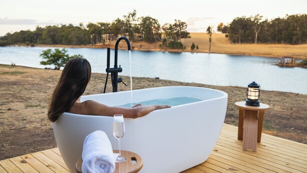 A person relaxing in a large, white bathtub with a view of a lake and trees. - Tiny Matilda Summary: (Bridgetown)