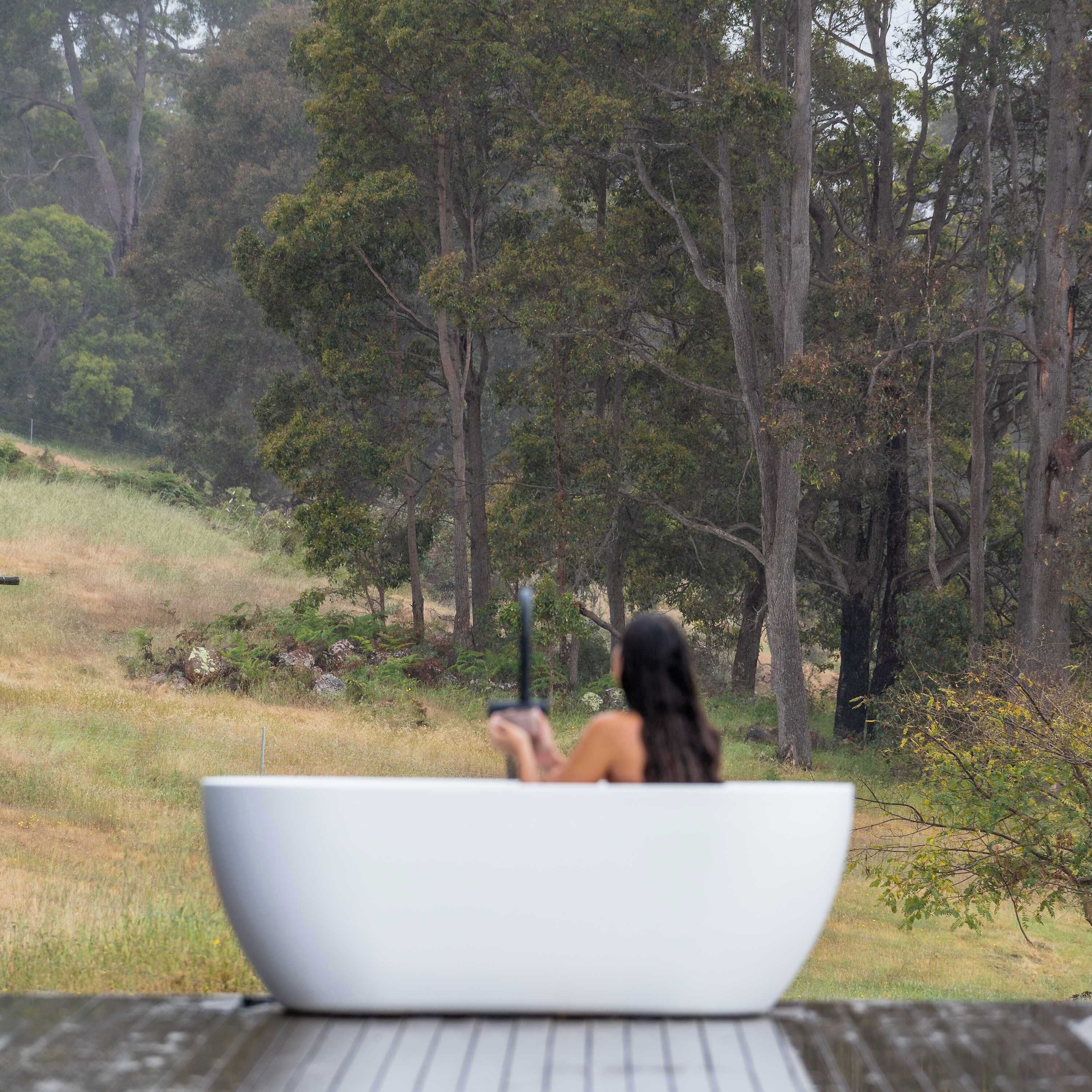 Une personne dans une baignoire blanche, à l’extérieur, tenant un téléphone intelligent, entourée d’arbres et d’herbe.