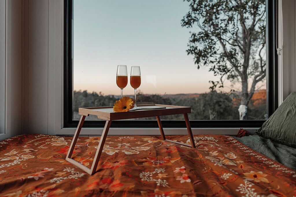 A bed with a floral bedspread, a wooden bedside table with two glasses of amber liquid, and a single flower.