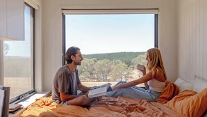 A bedroom with a large window, a bed with orange bedding, and a bedside table with books. - Tiny Rosie Summary:<br>S (Mount Crawford)