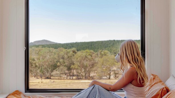A person sitting on a bed with a view of a forest and hills through a window. - Tiny Rosie Summary:<br>S (Mount Crawford)