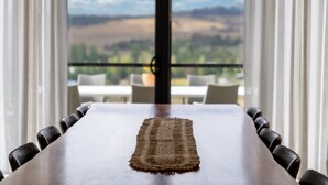 Una mesa de comedor con un mantel tejido, rodeada de sillas, con vista a un balcón y paisaje a través de grandes ventanales.