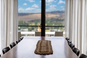 A dining table with a woven placemat, surrounded by chairs, with a view of a balcony and landscape through large windows.