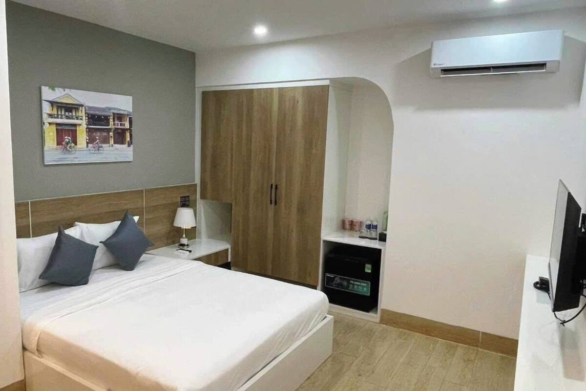 A hotel room with a bed, a wardrobe, a desk with a lamp, a TV, and an air conditioning unit.