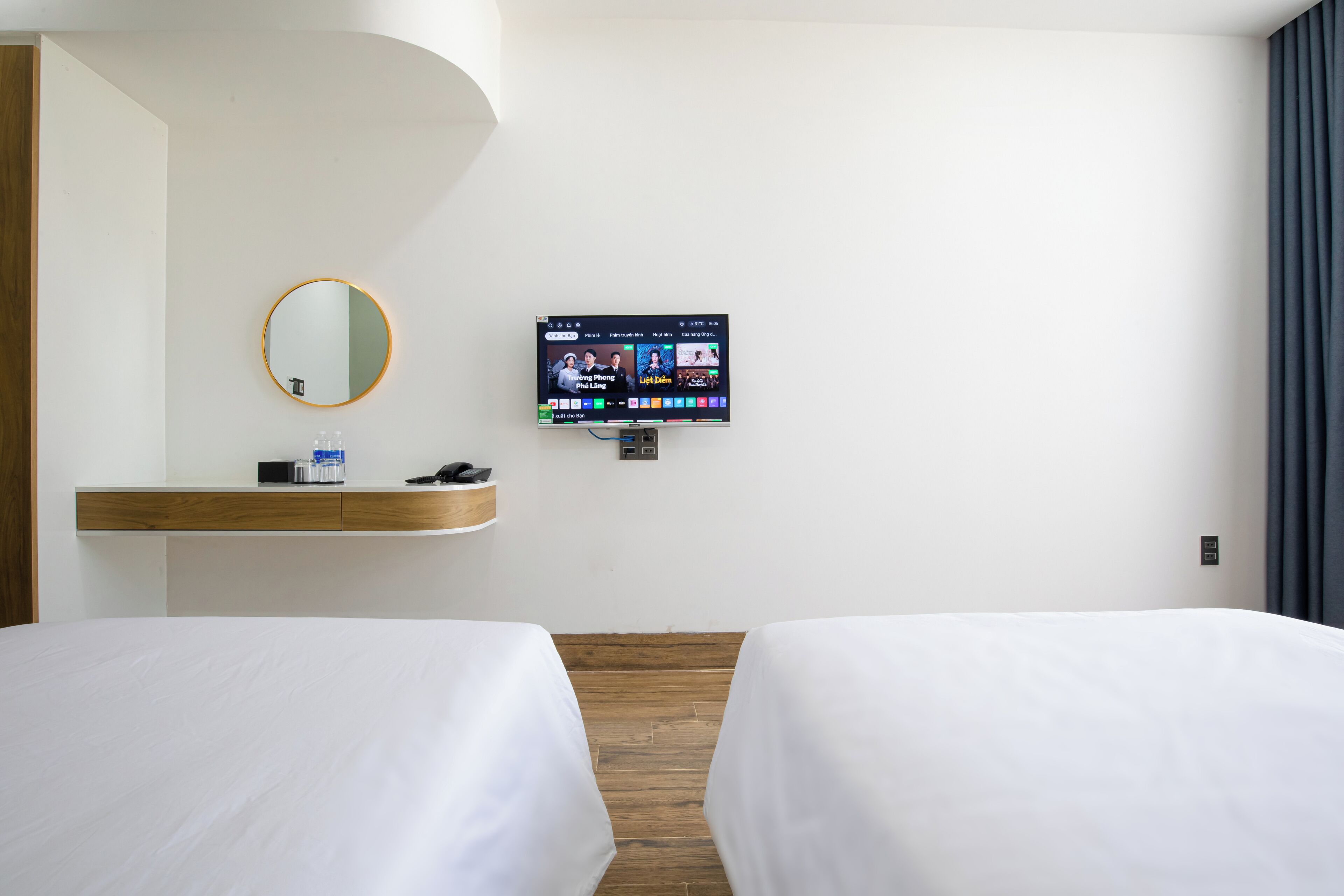 A hotel room with two beds, a TV mounted on the wall, a bedside table with a phone and a bottle, and a mirror on the wall.