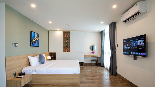 A modern hotel room with a bed, bedside table, desk, chair, TV, air conditioning unit, and a wall-mounted shelf with decorative items. - FIRST Studio Hotel Da Nang (Da Nang)