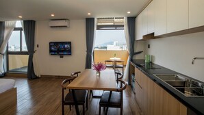 A modern hotel room with a wooden dining table, a flat-screen TV mounted on the wall, a kitchen area with a sink, and large windows offering a city view. - FIRST Studio Hotel Da Nang (Da Nang)