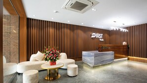 A modern reception area with a marble reception desk, a sofa, ottomans, and a flower arrangement. - FIRST Studio Hotel Da Nang (Da Nang)