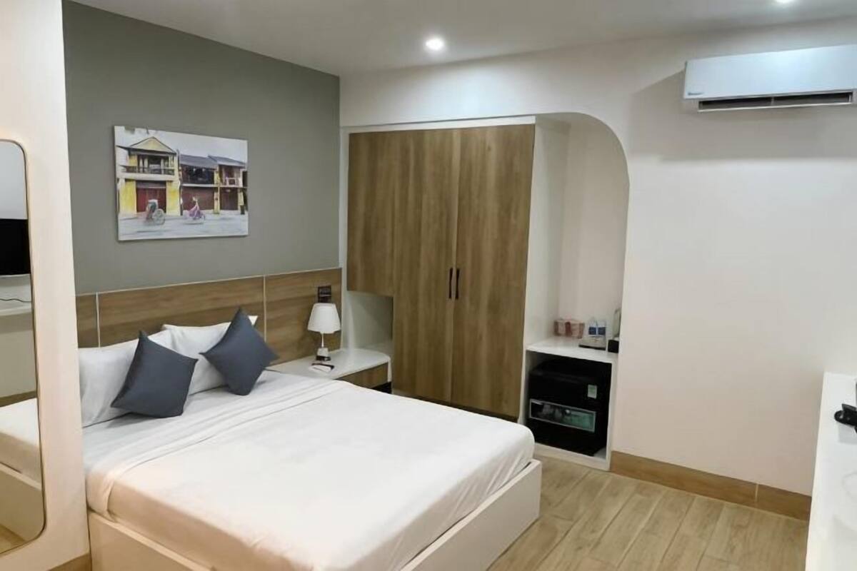 A hotel room with a bed, bedside table, lamp, wardrobe, and a wall-mounted air conditioner.