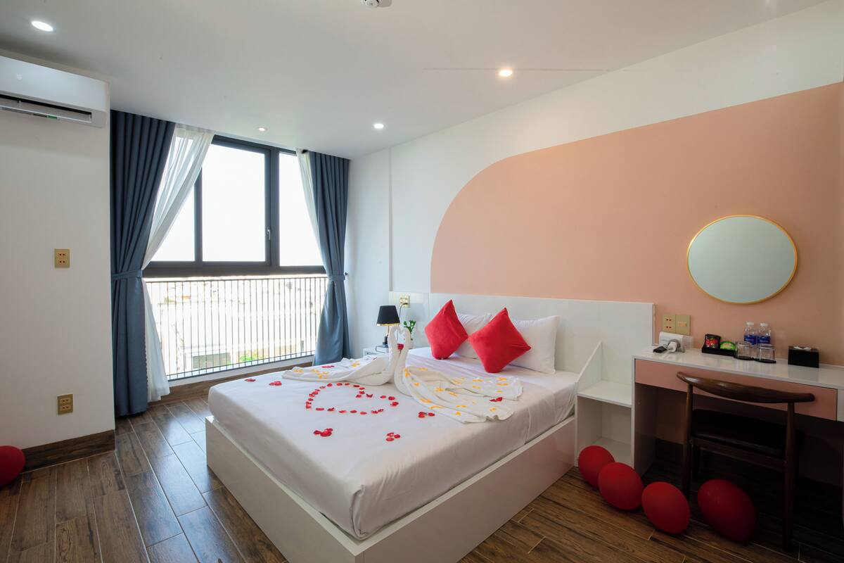 A hotel room with a large bed, a desk, a chair, a round mirror, and a balcony with curtains.