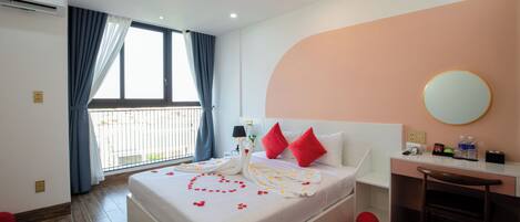 A hotel room with a large bed, a desk, a chair, a round mirror, and a balcony with curtains.