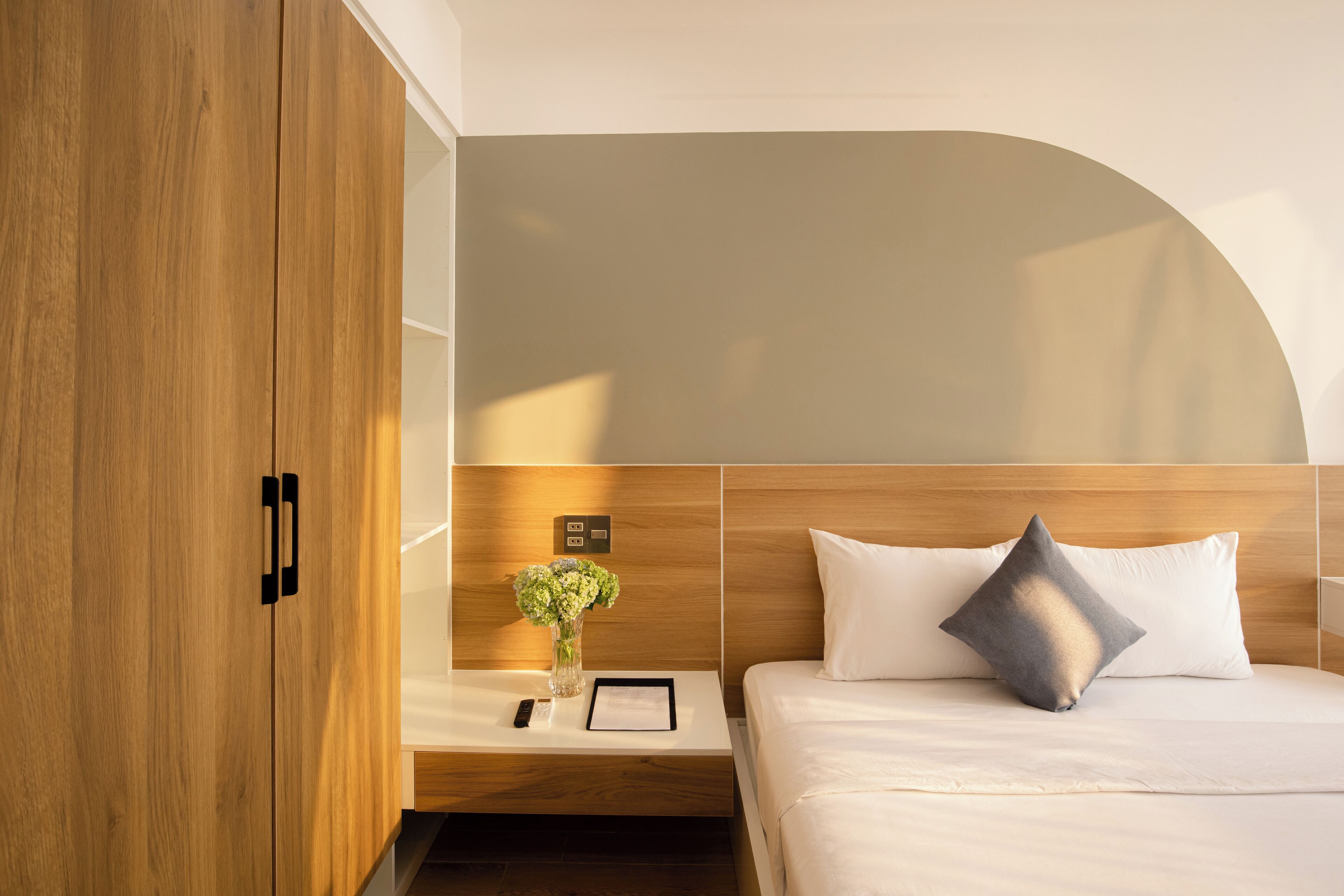 A modern hotel room with a wooden wardrobe, a bedside table with a vase of flowers, a bedside lamp, and a bed with white bedding and a gray pillow.
