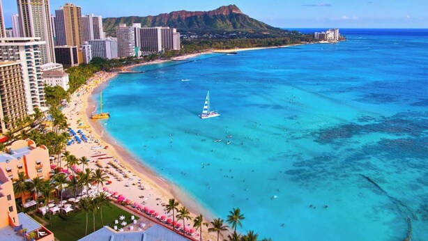 A coastal cityscape with a beach, clear blue waters, and a sailboat. - Waikiki Banyan condo w/ ocean & skyline views (Honolulu)