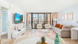 A modern living room with a sofa, chairs, a dining table with a pineapple and bottle, and a large window with a city view. - Waikiki Banyan condo w/ ocean & skyline views (Honolulu)