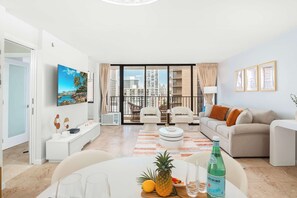 A modern living room with a sofa, chairs, a dining table with a pineapple and bottle, and a large window with a city view.