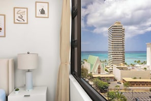 A hotel room with a view of a tall building, a lamp, and framed artwork on the wall.