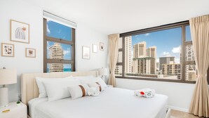 A hotel room with a bed, a nightstand, a lamp, a window with curtains, and framed pictures on the wall. - Waikiki Banyan condo w/ ocean & skyline views (Honolulu)