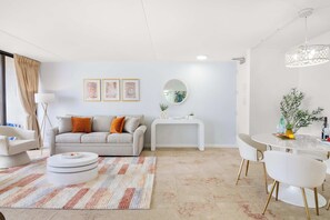 A modern living room with a sofa, a coffee table, a dining table with chairs, a lamp, and wall art.