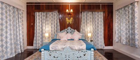 Un dormitorio con una cama grande, paneles de madera en las paredes, cortinas estampadas y un ventilador de techo.