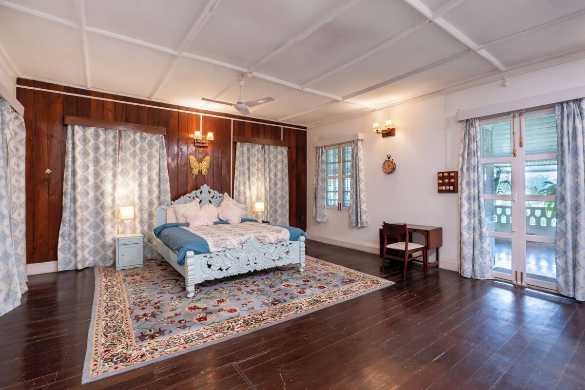 Ein Schlafzimmer mit einem großen Bett, Holzwänden, einem gemusterten Teppich, einem Nachttisch, einem Sessel und einem Fenster mit Vorhängen.