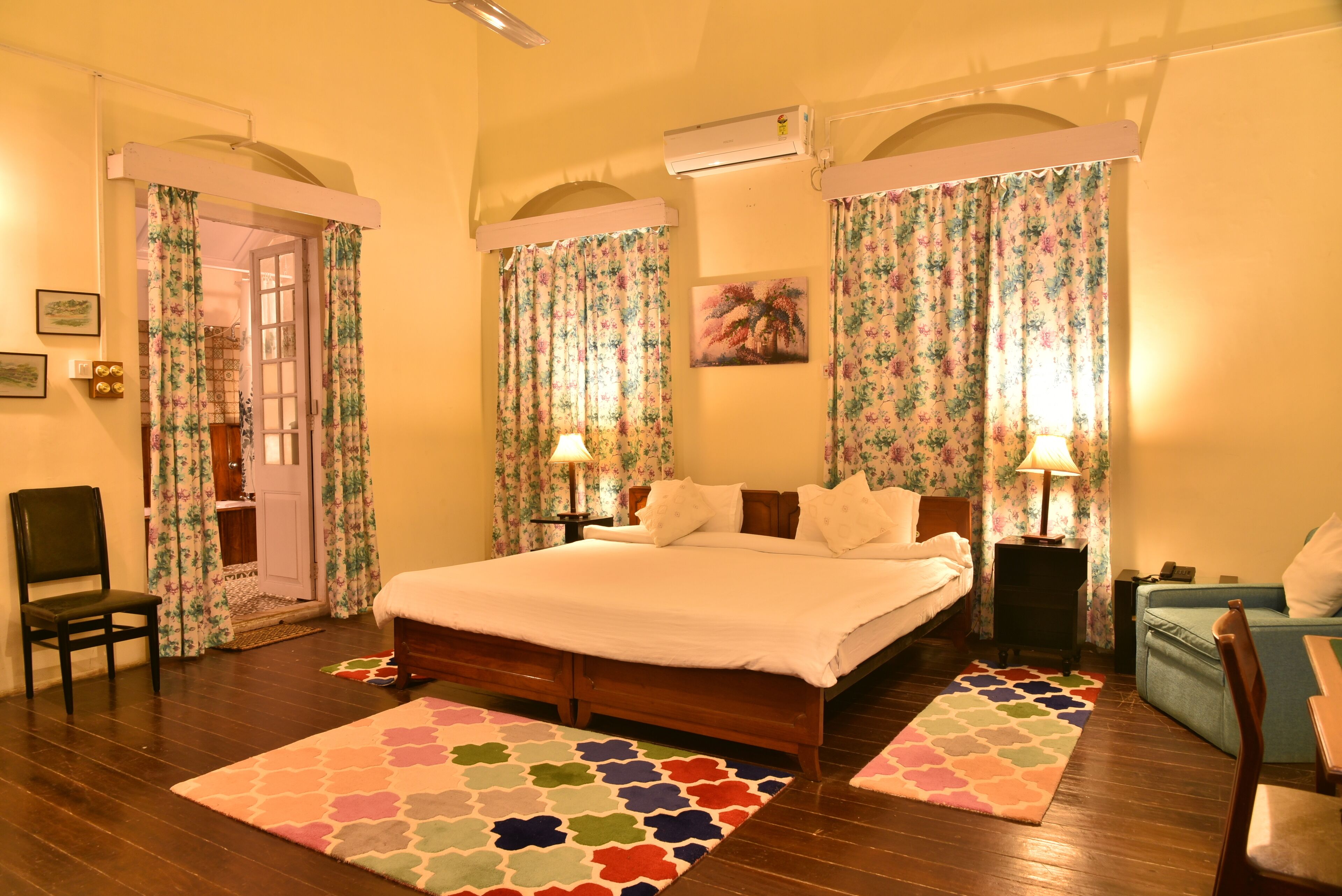 A bedroom with a wooden bed, two bedside tables with lamps, a chair, a sofa, and a door leading to another room.