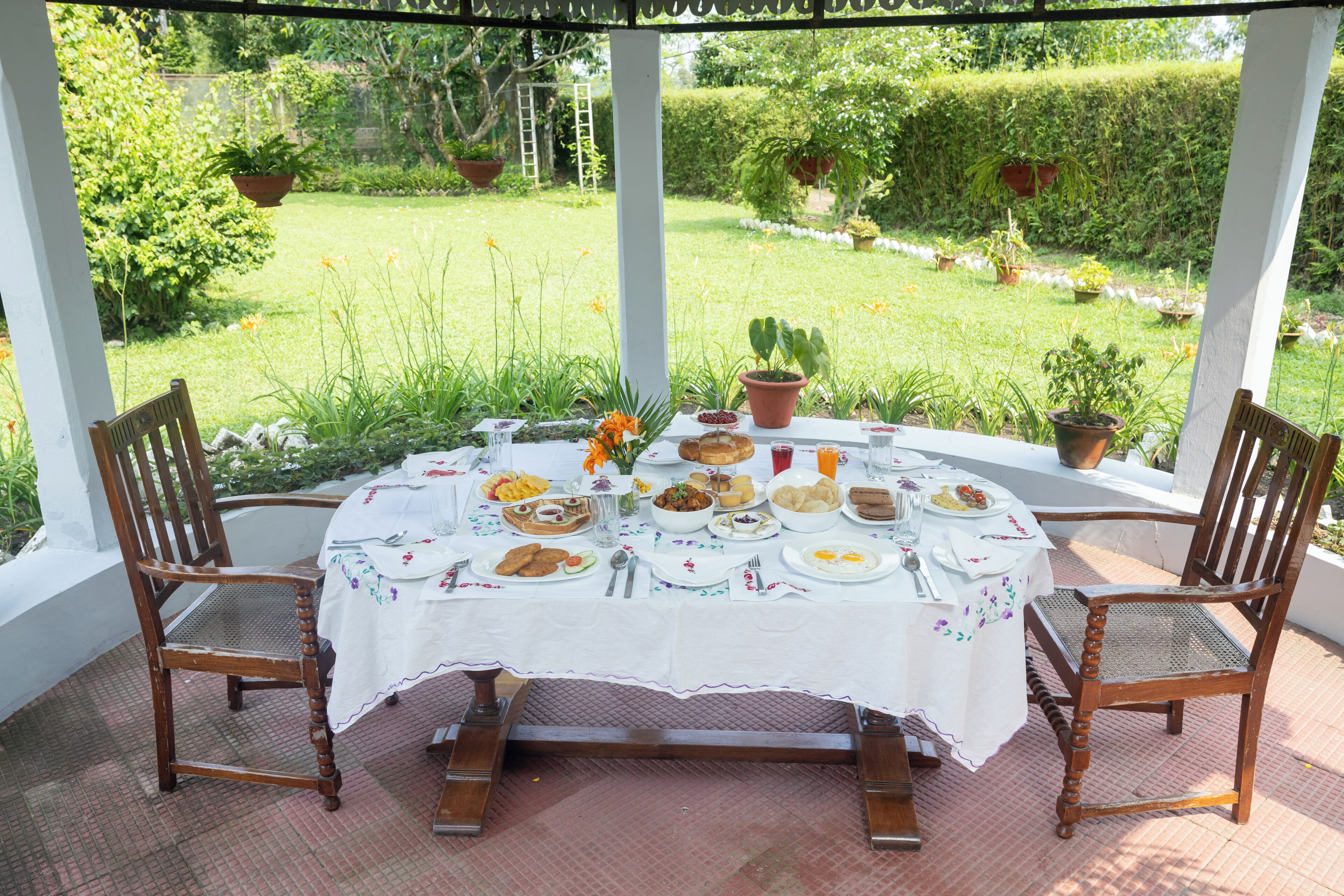 Un patio cubierto con una mesa redonda dispuesta para una comida, rodeado de sillas de madera, con vistas a un jardín con plantas en macetas y un seto.