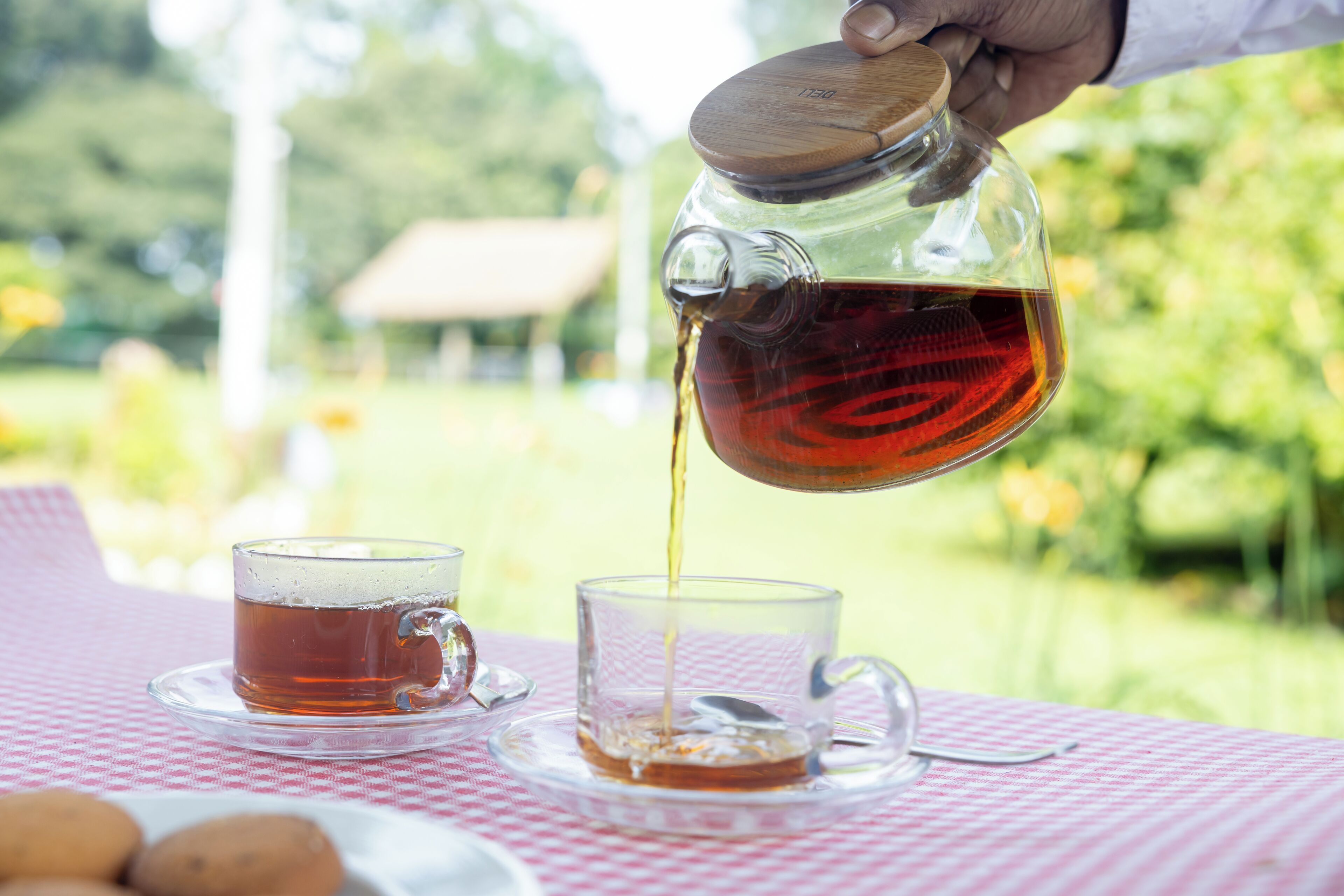 Une main verse du thé dans une tasse en verre posée sur une nappe à carreaux.