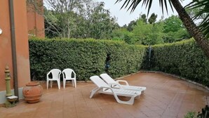 A patio with a white lounge chair, two white plastic chairs, a large terracotta pot, and a small fountain. - In the Green near the Sea (Orbetello)