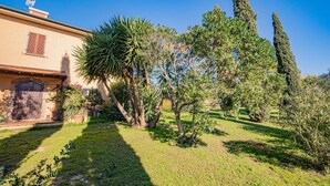 Un jardin bien entretenu, agrémenté de divers arbres et arbustes, se trouve devant un bâtiment doté d’un toit en tuiles et d’un petit balcon.