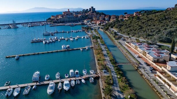A coastal marina with boats docked, a narrow road alongside, and buildings on a hillside. - View of the harbour (Orbetello)
