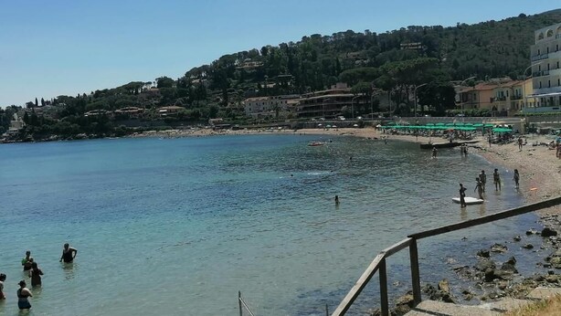 Zona costera con agua cristalina, una playa de arena y edificios en una colina.