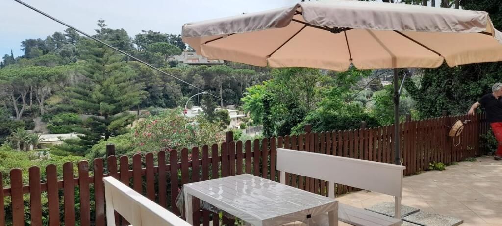 A patio with a table and chair under a beige umbrella, surrounded by a wooden fence and greenery.