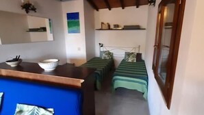 Una habitación con techo de madera, una cama con colcha verde, una mesita con un cuenco, una silla azul y una ventana con marco de madera.
