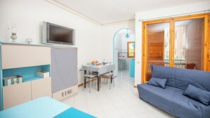 A compact living space with a dining area, a sofa, a TV, and a kitchenette. - The sea is close at hand. (Orbetello)