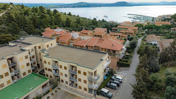 A residential area with multi-story buildings, a parking lot, and a view of a lake with boats. - The sea is close at hand. (Orbetello)