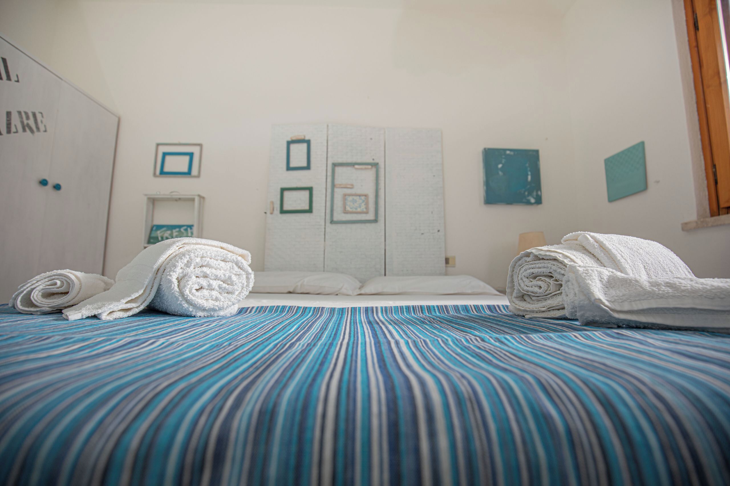 Un lit bien fait, avec des serviettes pliées, une couverture rayée bleue et blanche et une armoire blanche dont la porte porte une inscription.