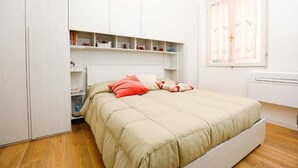 A bedroom with a bed, white wardrobe, shelves, and a window with curtains.