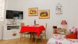 A small, well-lit room with a dining table, chairs, a TV, and a bed with floral bedding.