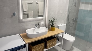 A modern bathroom with a large mirror, white sink, and a shower area. - Modern Seaside Home with Private Patio + Parking - Blue Zone Homes (Olimpiaki Akti)