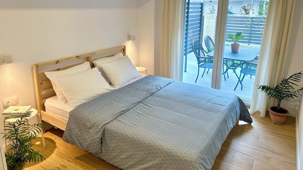 A bedroom with a wooden bed, a bedside table, a window with curtains, and a view of an outdoor patio with chairs and a table. - Modern Seaside Home with Private Patio + Parking - Blue Zone Homes (Olimpiaki Akti)