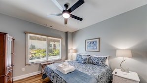 A bedroom with a bed, bedside table, lamp, window with blinds, ceiling fan, and a framed picture on the wall. - Alpine Village Condo E| Location, Location, Location, Across From RIver and Tubing!! (Helen)