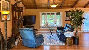 A cozy living room with a wooden ceiling, a fireplace, a television, a blue armchair, a leather sofa, and a potted plant.