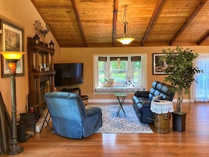 A cozy living room with a wooden ceiling, a fireplace, a television, a blue armchair, a leather sofa, and a potted plant.