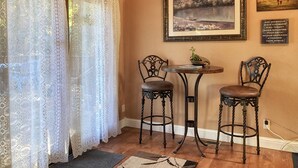 A room with a wooden ceiling, a white lace curtain, two ornate chairs, a small round table with a plant, and a framed picture on the wall.