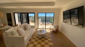 Un salon spacieux avec un canapé blanc, une table basse ronde et un fauteuil en osier. De grandes fenêtres offrent une vue sur le jardin.
