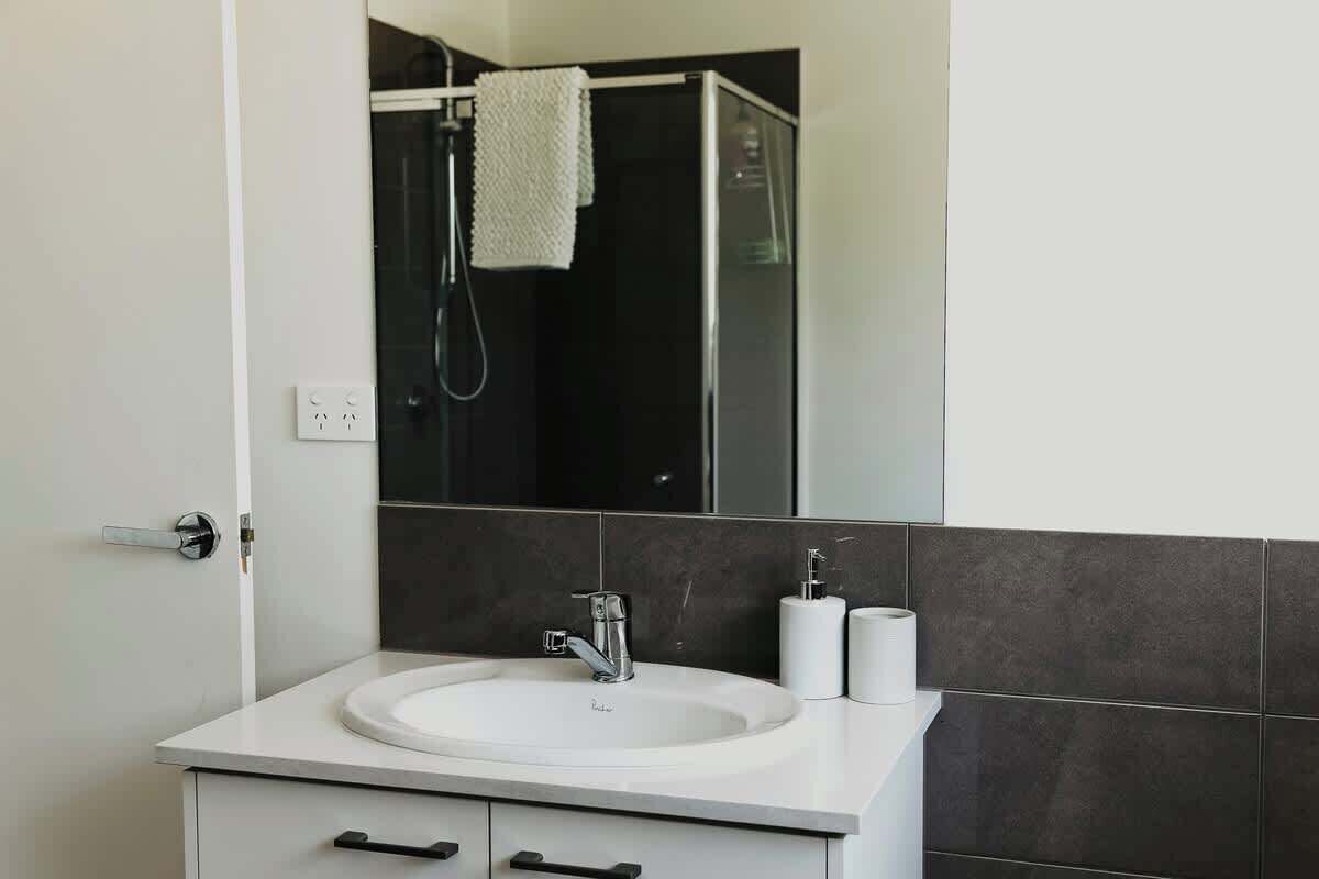 Un baño moderno con un lavabo blanco, paredes cubiertas de baldosas oscuras, un espejo grande y una zona de ducha.