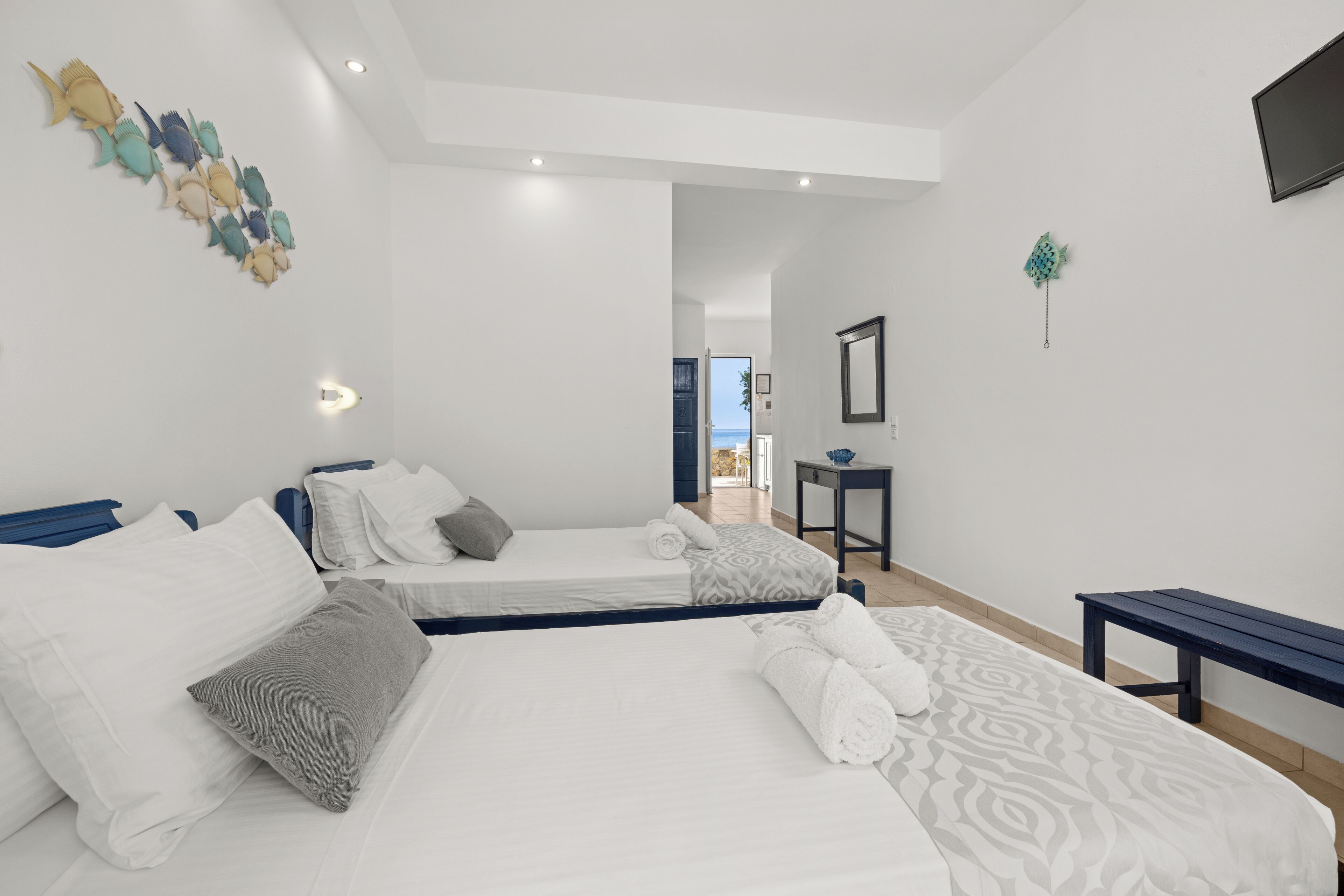 A modern hotel room with two beds, a small table, a bench, and a view of the sea through an open door.